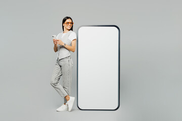 Full body side view young latin woman she wears white t-shirt gray vest glasses big huge blank screen mobile cell phone with mockup workspace area using smartphone isolated on plain grey background.