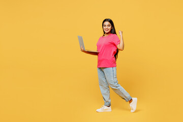 Fototapeta na wymiar Full body side view young IT Indian woman wear pink t-shirt casual clothes hold use work on laptop pc computer do winner gesture isolated on plain yellow background studio portrait. Lifestyle concept.