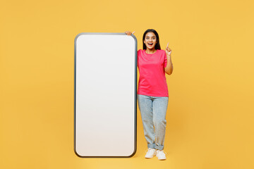 Full body young insighted Indian woman wear pink t-shirt casual clothes big huge blank screen mobile cell phone smartphone with area point index finger up isolated on plain yellow background studio.