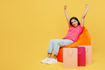Young happy woman wearing casual clothes sit in bag chair near paper package bags after shopping do winner gesture isolated on plain yellow color background studio. Black Friday sale buy day concept.