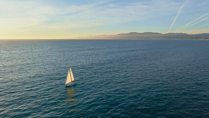 Calm sea at sunset with a single sailboat gliding across the water near the coastline