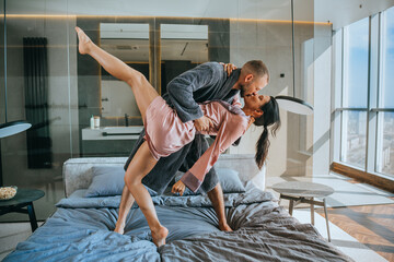 Young man in robe kissing girlfriend at hotel room, dancing on bed enjoying vacations on summer....