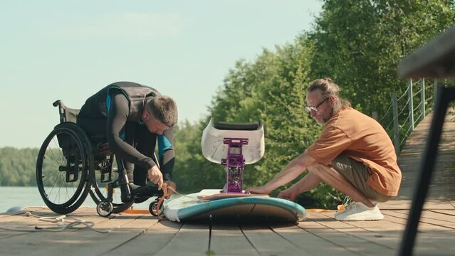 Wheelchaired man in wetsuit and adaptive sup surfing instructor getting Supboard ready for surfing on wooden pier on warm sunny dayWheelchaired man in wetsuit and adaptive sup surfing instructor getti