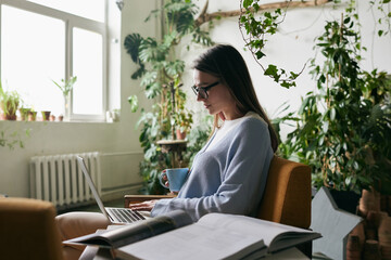 A portrait of a girl in glasses with coffee in her hand and laptop on her knees in profile