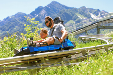 father and child having ride on summer toboggan called Rodelbahn rushing down the track . Beautiful mountains on background - obrazy, fototapety, plakaty