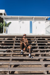 Chico joven musculoso y tatuado posando sin camiseta y en bañador frente a chiringuito de playa