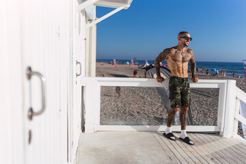Naklejka premium Chico joven musculoso y tatuado posando sin camiseta y en bañador frente a chiringuito de playa