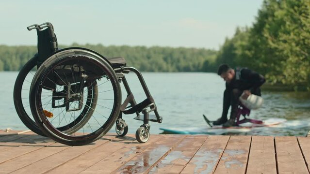 Young athletic man with disability in wetsuit paddling on sit down supboard in river with selective focus on wheelchair under sunlight at wooden pier