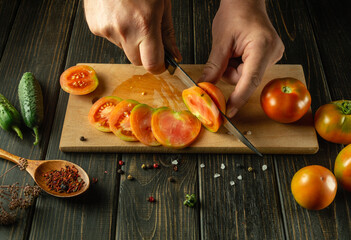 Hands of a man with a knife slicing a tomato on a cutting board. The concept of cooking vegetable salad with cucumbers and tomatoes at home