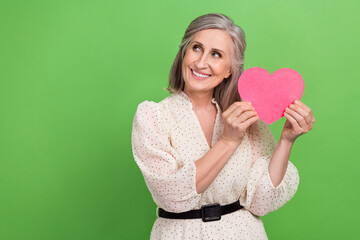 Photo portrait of lovely retired lady hold pink heart card look empty space wear trendy white outfit isolated on green color background