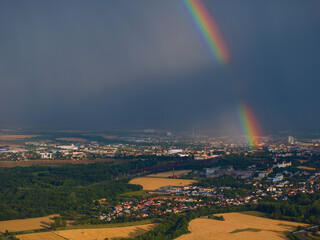  Aerial view of a massive rainbow arc ending in the city and a receding summer storm over the cultural landscape with the city in the background. Dramatic sudden summer storms.