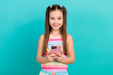 Portrait of little positive girl toothy smile hold use smart phone chatting isolated on turquoise...