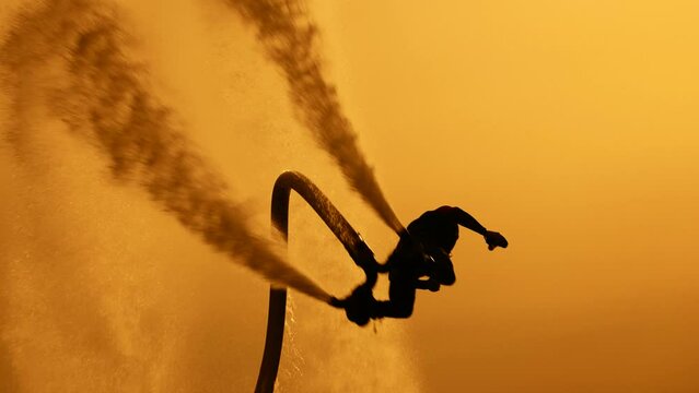 The silhouette of a flyboarder against the sun and the sunset sky. The athlete performs flips and other tricks over the water. The freedom and agility of an extreme athlete.