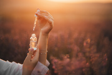 Close up female hands holding essential oil in a bottle with pipette in lavender field at sunset.