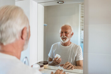 Men's personal care. Bearded senior man puts cream on his face in the bathroom.