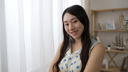 shoulder shot portrait of an attractive Asian woman look at the camera with smile near the window at home during the day.