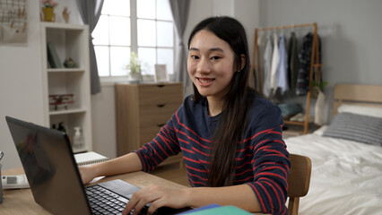 portrait of a cheerful Asian college girl looking at the camera with a smile while learning from home on the computer at desk in a modern bedroom.