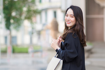 Portrait of beautiful long haired young asian business woman in black coat carrying walking bag smiling in good mood in the city outdoors.