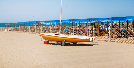 Small boat - sailboat , light brown Dark orange color. He stands on a sandy beach, with blue umbrellas in the background. Format: panorama