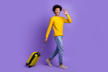 Full body photo of friendly young student guy waves palm hello sign walking with luggage traveling isolated on violet color background