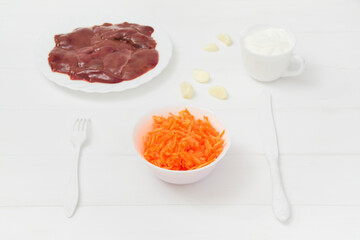 Fresh raw chicken liver and grated carrots in a plate and garlic cloves, Sour cream gravy boat. White wooden background. Light key.