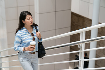 stressed asian Korean business lady carrying morning coffee and walking upstairs in white modern...