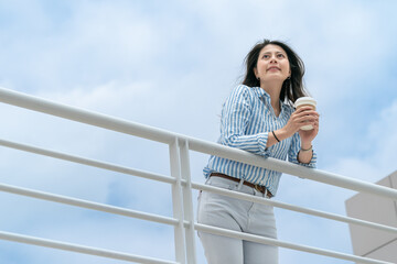 view from below of smiling asian korean businesswoman relaxing on railing and looking up into sunny blue sky with takeout coffee cup in hand