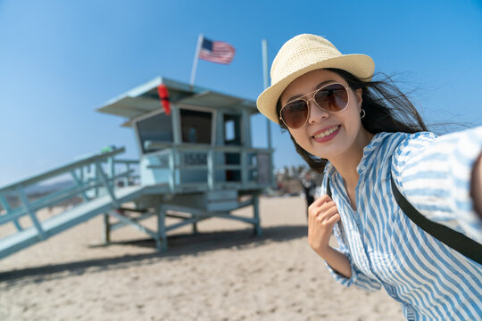 happy asian Korean lady tourist wearing hat and sunglasses smiling at camera taking selfie picture with Lifeguard Tower at back on santa monica beach