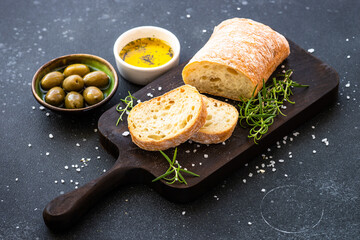 Ciabatta bread on wooden board with olive oil, olives and herbs on black. Mediterranean food.