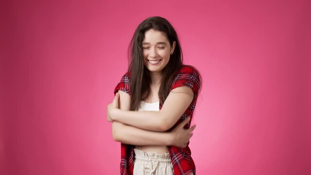 Contented casually dressed young smiling woman with long dark hair hugging herself with both arms while smiling peacefully against pink background