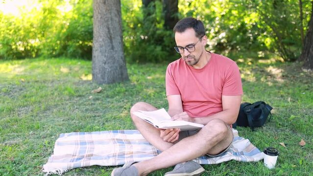A man reads a book sitting on a blanket on the grass in a park on a summer day.