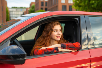 With a radiant smile, a young woman with ginger hair sits in the red car, looking through the open window.