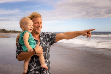 Smiling father and happy baby son spending time on sandy beach. Summer vacation. Family relationships. Happy childhood in Asia. Caucasian man pointing towards the horizon. Seminyak beach, Bali