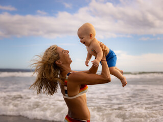 Mother holding, smiling, laughing and lifting her infant baby boy son high in the air on sandy beach. Family having fun together outdoors. Motherhood concept. Seminyak beach, Bali