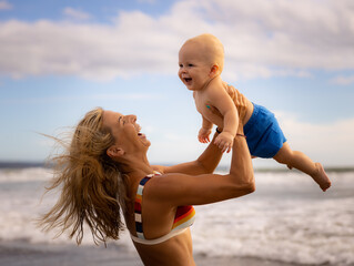 Mother holding and lifting her infant baby boy high in the air on beach. Positive emotions. Mum and son laughing and smiling. Blue sky with white clouds. Summer vacation. Seminyak beach, Bali