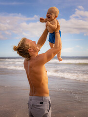 Father holding and lifting his infant baby boy high in the air on beach. Positive emotions. Dad and son laughing and smiling. Blue sky with white clouds. Summer vacation. Seminyak beach, Bali