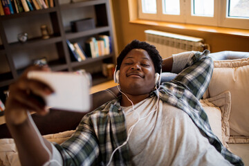 Young man of african ethnicity using a smart phonew while sitting on the couch and listening to music in the living room