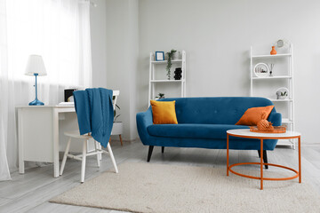 Interior of light living room with blue sofa, shelving units, workplace and coffee table