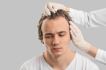 Doctor examining young man with hair loss problem on grey background, closeup