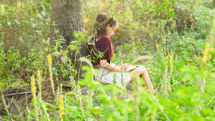 Naklejka na ściany i meble Young woman drawing in a notebook, sitting by a tree