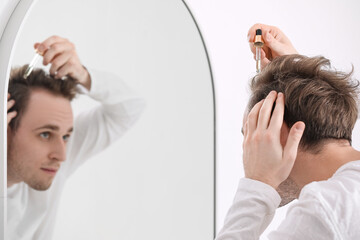 Young man using serum for hair growth near mirror in bathroom, closeup
