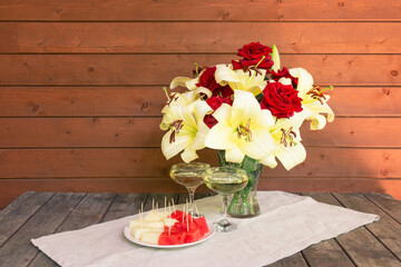 White lilies and red roses, glasses of champagne and plate with watermelon and melon on rustic wooden table.