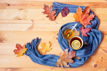 Mug of hot tea, honey, autumn leaves and scarf on wooden background. Top view, copy space.