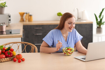 Obraz na płótnie Canvas Young woman with laptop eating vegetable salad in kitchen