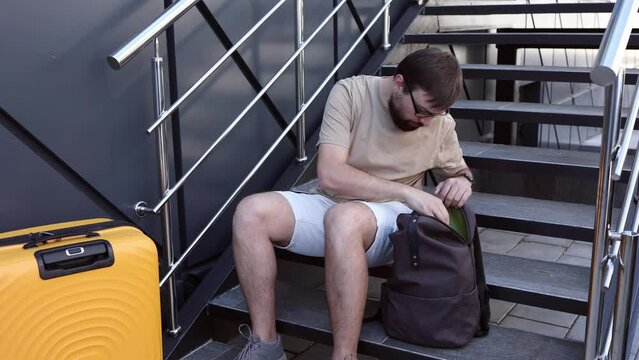 Man sits with a beard, glasses looking in his backpack for tickets before traveling. Concept: Trip, Travel, Plane, Ticket Search, Orange Suitcase, Day One, Migration, Immigration, Passport