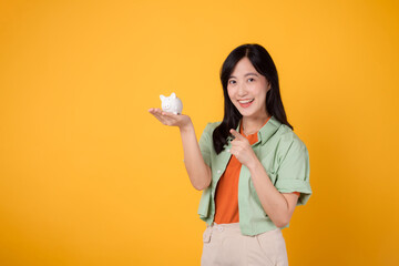 young Asian woman in her 30s, elegantly dressed in orange shirt and green jumper, revealing piggy bank while pointing finger to free copy space on yellow background. Financial money concept.