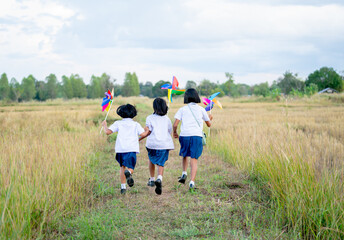 Back of little Asian girls hold small windmill and look fun to run together in walkway of rice field.