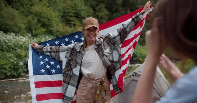First person method a girl photographs her friend who is standing with the flag of the United States of America in a cap against the backdrop of a green forest