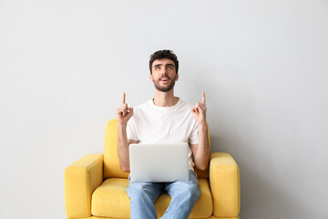 Young man with laptop pointing at something in armchair on light background