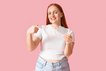 Young woman pointing at contraceptive pills on pink background